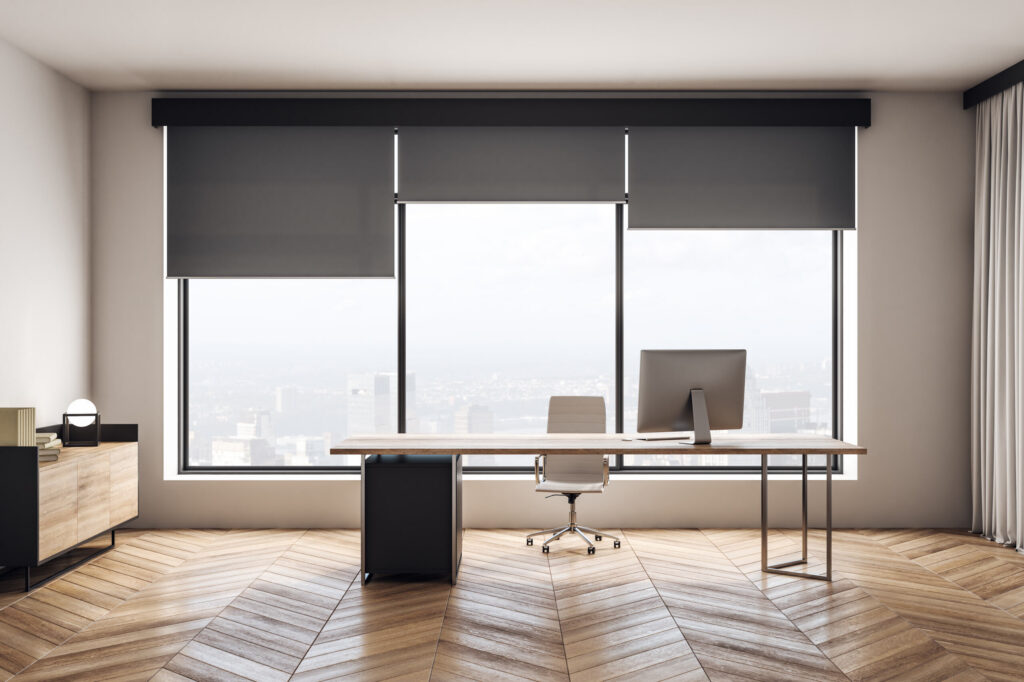 Modern office interior featuring large windows with grey roller blinds, wooden desk, computer, and city view in the background.